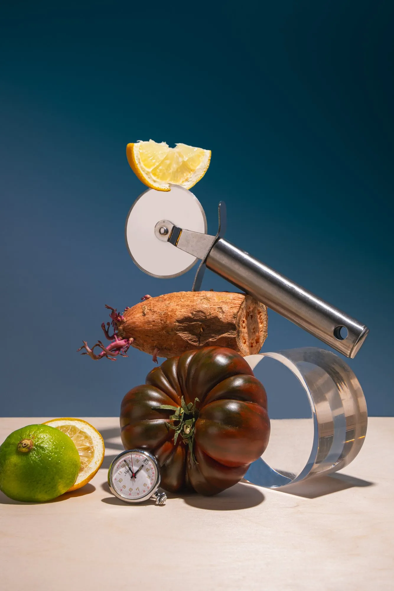 A pizza cutter balanced on a sweet potato and a tomato, with lemons and a stopwatch on blue